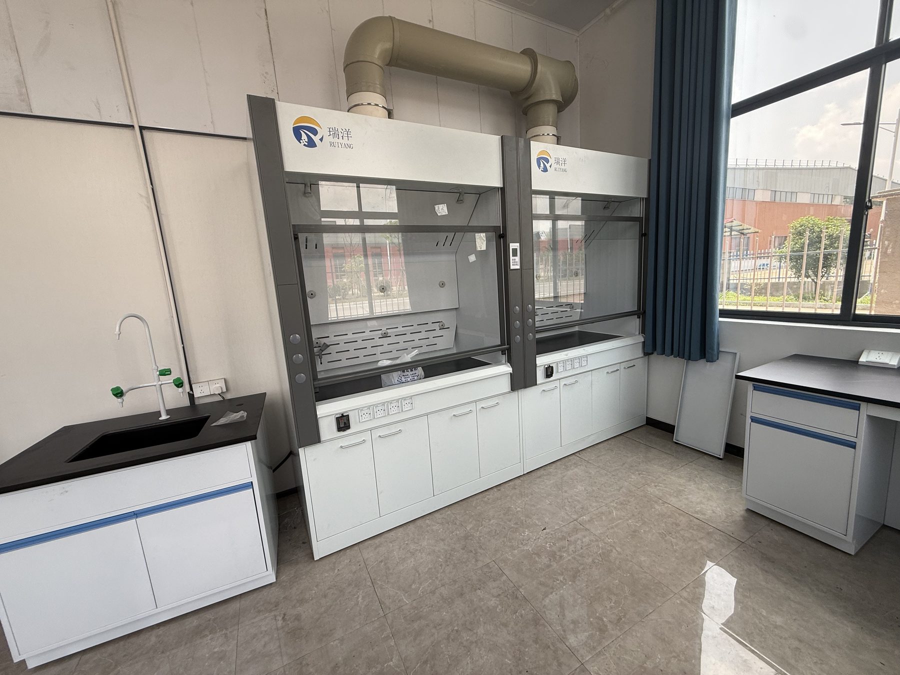 Installed laboratory fume hood connected to an overhead exhaust duct beside a wall bench